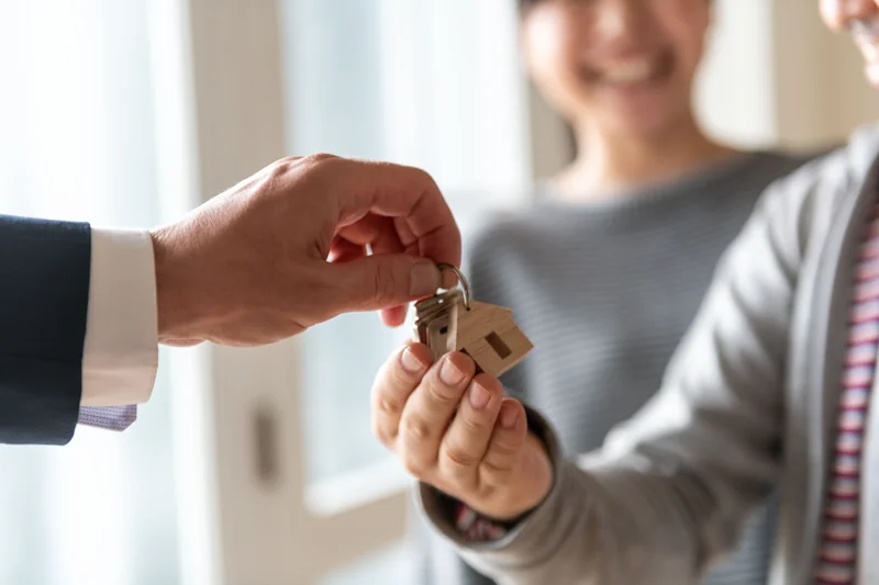 Receiving the Keys to Your New Home | Buyer's Guide Real estate agent handing house keys with a wooden house keychain to a smiling new homeowner