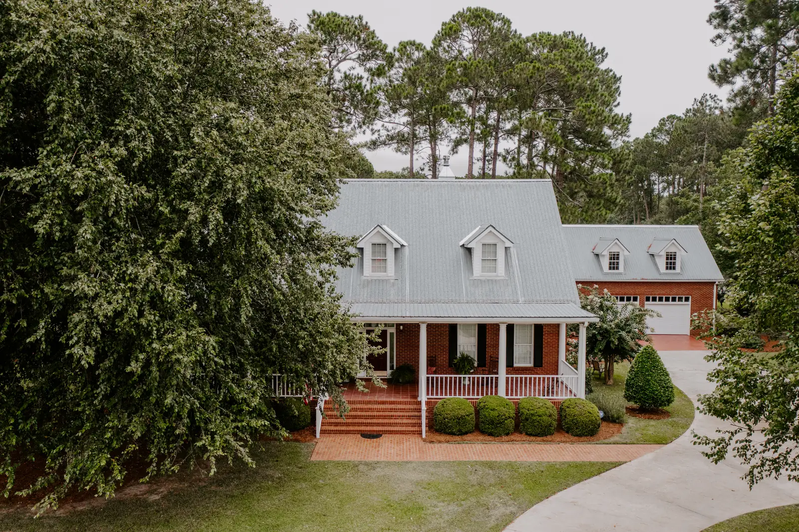 Two-story cream brick and siding home with dark shutters, an attached two-car garage, trimmed hedges, and a green front lawn under mature shade trees