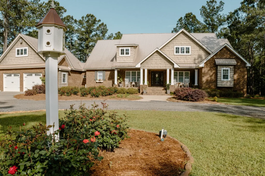 Homes for Sale in Hazlehurst, Georgia Single-story red brick home with cream gable siding, diamond-pane windows, and colorful zinnia and marigold flower beds under tall shade trees