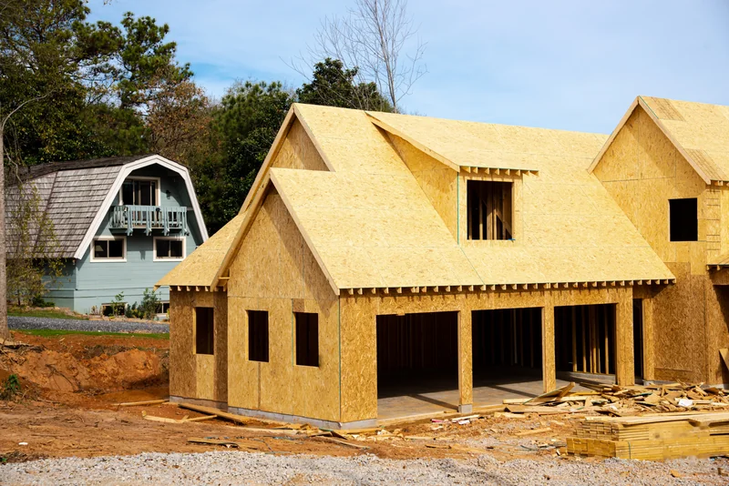 New home framed in OSB sheathing on a cleared red-clay lot beside an existing house, showing a building site under construction in Georgia